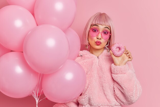 Mulher asiática adorável romântica mantém os lábios dobrados tem cabelo bob rosa vestido com casaco de pele de inverno contém deliciosa rosquinha vitrificada e balões de hélio inflados comemora aniversário na festa com amigos.