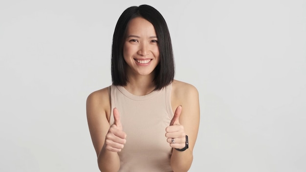 Mulher asiática alegre mantendo os polegares para cima isolados em fundo branco Mulher asiática parecendo feliz aprovando uma boa ideia Gosto