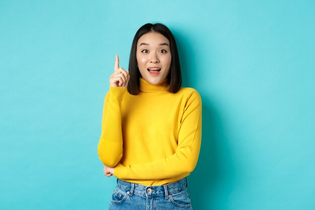 Mulher asiática animada com cabelo escuro curto, lançando uma ideia, levantando o dedo em gesto de eureca e sorrindo, em pé sobre um fundo azul
