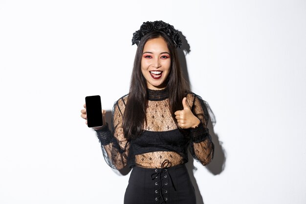 Foto grátis mulher asiática animada e satisfeita com fantasia de halloween, mostrando o polegar para cima em aprovação e demonstrando a tela do celular, em pé sobre uma parede branca