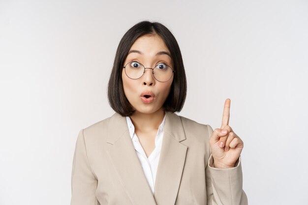 Mulher asiática animada em óculos, levantando o sinal de eureka do dedo tem uma ideia sobre fundo branco Copiar espaço