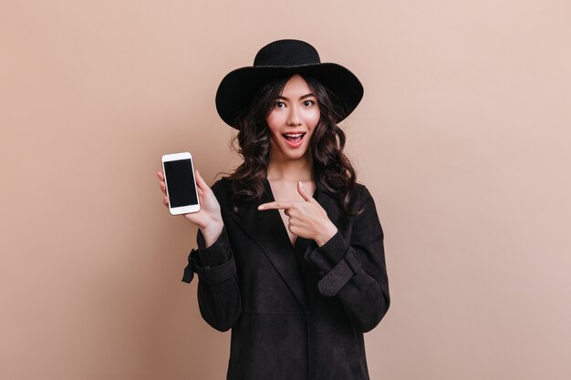 Mulher asiática, apontando com o dedo para smartphone com tela em branco. Foto de estúdio de emocional dama coreana com casaco preto.