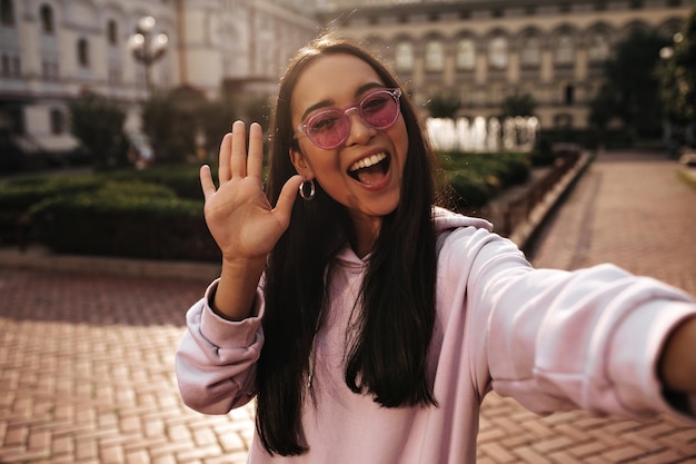 Mulher asiática atraente com capuz rosa e óculos de sol sorri acena com a mão em saudação e tira selfie do lado de fora
