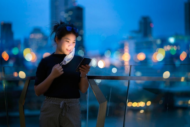 Mulher asiática atraente usa máscara protetora mão usar comunicação de tecnologia de smartphone com fundo de conceito de ideias de negócios de fundo de construção urbana luz bokeh