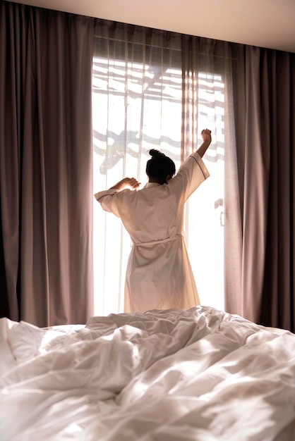 Mulher asiática bonita de vestido branco que estica a manhã acorda frescura e momento de fim de semana despreocupado no quarto interior aconchegante com cortina branca e conceito de casa de estilo de vida de fundo de brilho de sol