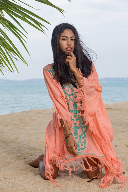 Mulher asiática bronzeada sexy incrível posando na praia tropical do paraíso sob a árvore pam, sentado na areia branca, relaxando e curtindo as férias. Vestido Boho com bordado. Bali.