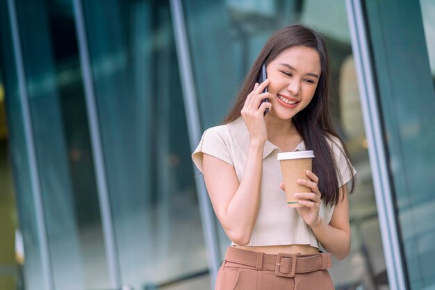 Mulher asiática casual relaxa programador nômade digital parece conversa no smartphone enquanto caminha na cidade com uma xícara de café segurando um sentimento positivo alegre e sorridente, reduzindo o estilo de vida