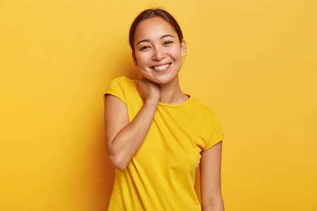 Mulher asiática charmosa tem expressão alegre, toca o pescoço e inclina a cabeça, tem conversa agradável com o interlocutor, ouve notícias agradáveis, usa camiseta casual vívida, isolada na parede amarela