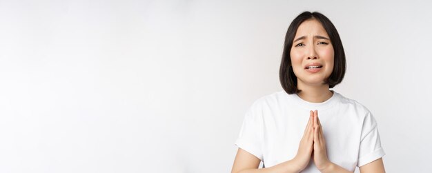 Mulher asiática chorando desesperada implorando por ajuda pedindo e dizendo por favor de pé na camiseta branca sobre fundo branco