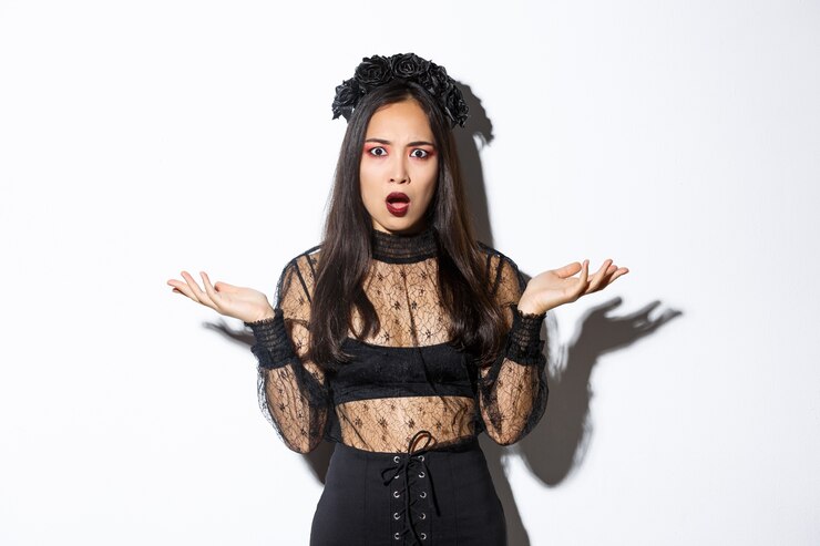 Foto grátis mulher asiática confusa com fantasia de halloween, parecendo sem noção, levantando as mãos para os lados e olhando perplexa para a câmera. mulher usando vestido de renda preta, sendo uma bruxa ou mágico em uma festa de doce ou travessura.