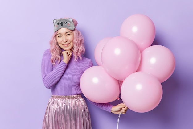 Mulher asiática de cabelo rosa satisfeita usa jumper de máscara de dormir e saia plissada segura balões inflados comemora aniversário isolado sobre fundo roxo. eventos especiais de pessoas e conceito de celebração