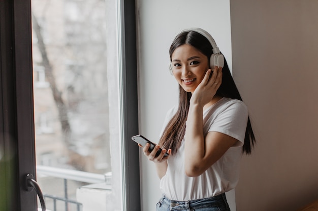 Foto grátis mulher asiática de olhos castanhos com top branco olha para a frente com um sorriso, segura o smartphone e coloca os fones de ouvido