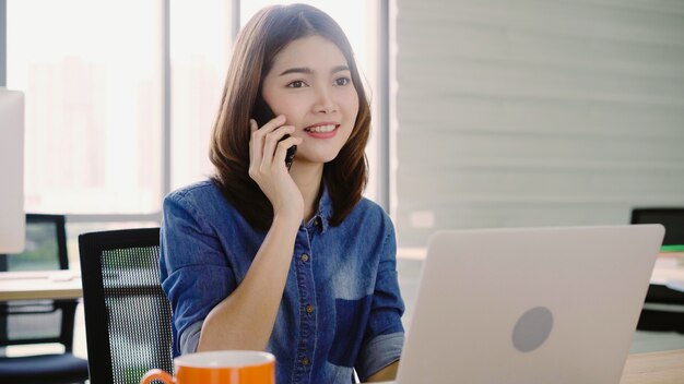 Mulher asiática de sorriso dos jovens bonitos que trabalha no portátil ao apreciar usando o smartphone no escritório.