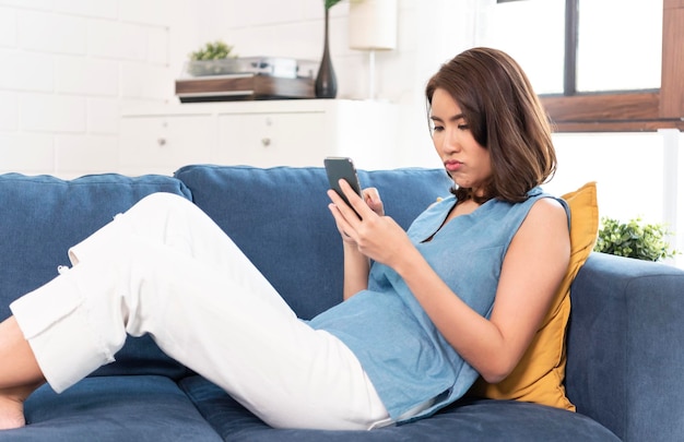 Mulher asiática deprimida usando smartphone, conversando em redes sociais no sofá em casa.