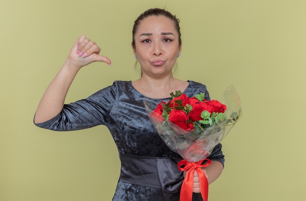 Mulher asiática descontente segurando um buquê de rosas vermelhas, olhando para a frente, mostrando o polegar para baixo, comemorando o dia internacional da mulher em pé sobre a parede verde