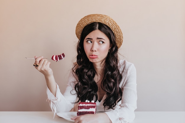 Mulher asiática engraçada no chapéu de palha comendo bolo. Modelo feminino chinês bonito apreciando a sobremesa.