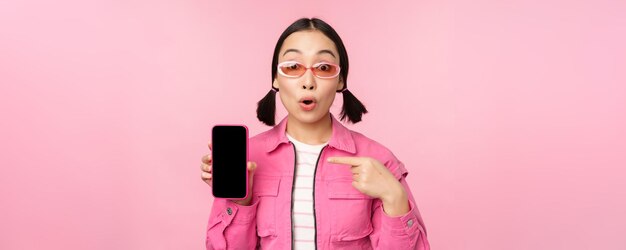 Mulher asiática entusiasmada em óculos de sol de roupas elegantes, apontando o dedo para a tela do celular, mostrando o aplicativo do smartphone sobre fundo rosa