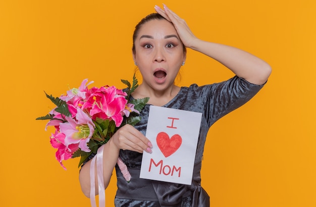 Mulher asiática espantada e surpresa segurando um cartão e um buquê de flores comemorando o dia das mães em pé sobre uma parede laranja