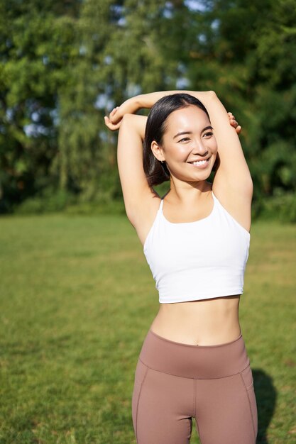 Mulher asiática esticando os braços fazendo exercícios físicos no parque sorrindo satisfeito aquecendo antes de correr