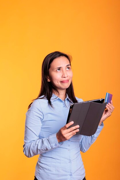 Mulher asiática fazendo compras on-line segurando um tablet, digitando informações de cartão de crédito ao fazer compras on-line. Cheerfu jovem adulto encomendando roupas da moda em estúdio sobre fundo amarelo