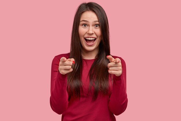 Mulher asiática feliz com longos cabelos escuros aponta para a câmera com os dois dedos indicadores, parabeniza amigo, escolhe diretamente você, usa roupas vermelhas