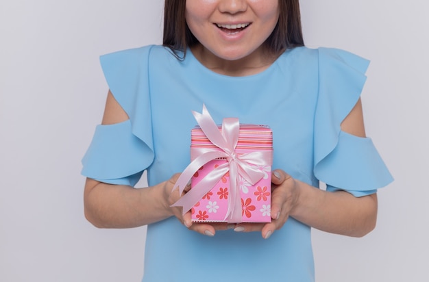 Mulher asiática feliz com vestido azul segurando um presente para comemorar o dia internacional da mulher em pé sobre uma parede branca