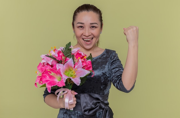 Mulher asiática feliz e animada segurando um buquê de flores com os punhos cerrados e sorrindo alegremente, comemorando o dia internacional da mulher em pé sobre a parede verde