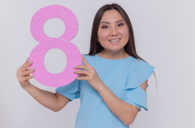 Mulher asiática feliz e positiva segurando o número oito feito de papelão, olhando para a frente, sorrindo alegremente, comemorando o dia internacional da mulher em pé sobre uma parede branca