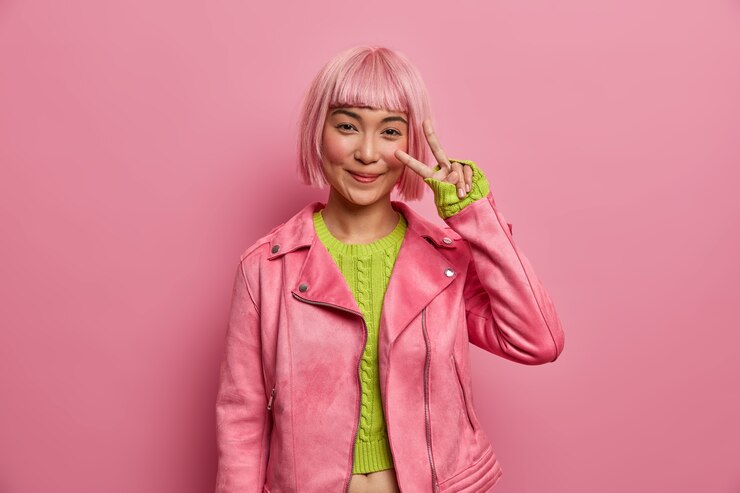 Foto grátis mulher asiática feliz mostra o sinal v, permanece tranquila e positiva, vestida com uma jaqueta rosa da moda, suéter verde, aproveita a vitória, gesticula ativamente