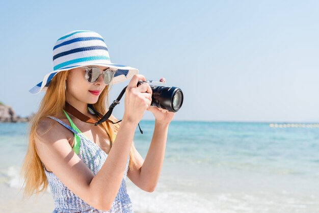 Mulher asiática feliz nas férias fotografando com uma câmera na praia com o horizonte no fundo.