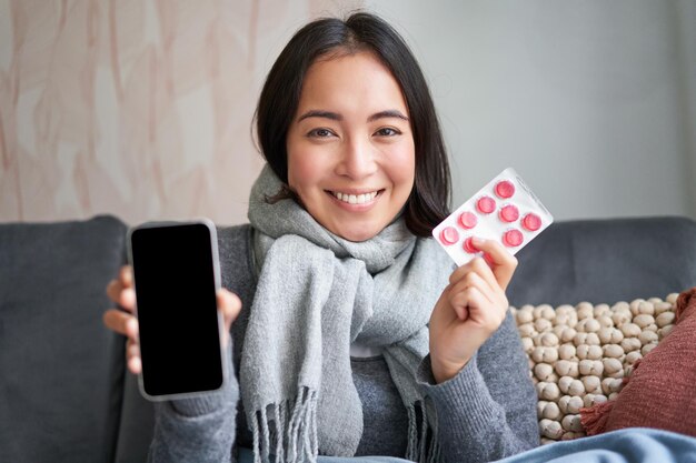 Mulher asiática feliz pegando resfriado mostrando a tela do smartphone e pílulas recomendando aplicativo médico on-line