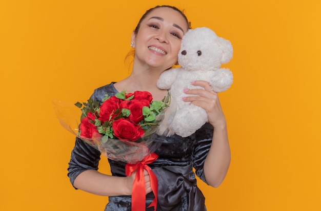 Mulher asiática feliz segurando um buquê de rosas vermelhas e ursinho de pelúcia como um presente, sorrindo alegremente, comemorando o dia das mães em pé sobre uma parede laranja