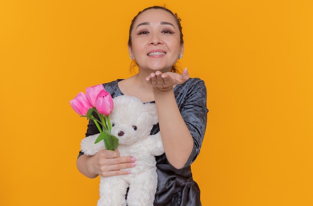 Mulher asiática feliz segurando um buquê de tulipas cor de rosa e ursinho de pelúcia mandando um beijo