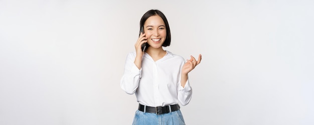 Mulher asiática feliz sorridente falando no smartphone com a vendedora do cliente de plantão segurando o celular e gesticulando em pé sobre fundo branco