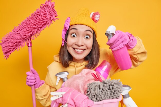 Mulher asiática feliz sorridente segurando frasco de spray e esfregão fica perto do cesto de roupa suja com frascos de produtos químicos e usa luvas de borracha e usa casa