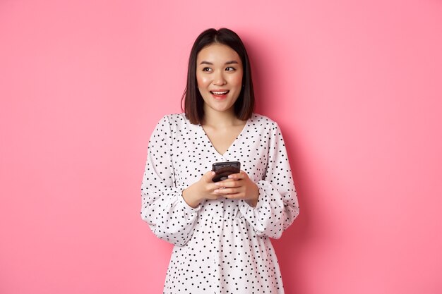 Mulher asiática fofa pensando e sorrindo, parecendo sonhadora enquanto envia uma mensagem no smartphone, navegando em lojas online, em pé sobre um fundo rosa