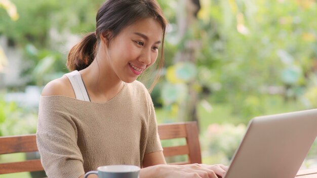 Mulher asiática freelance trabalhando em casa, negócios feminino trabalhando no laptop sentado na mesa no jardim de manhã. Mulheres do estilo de vida que trabalham em casa o conceito.