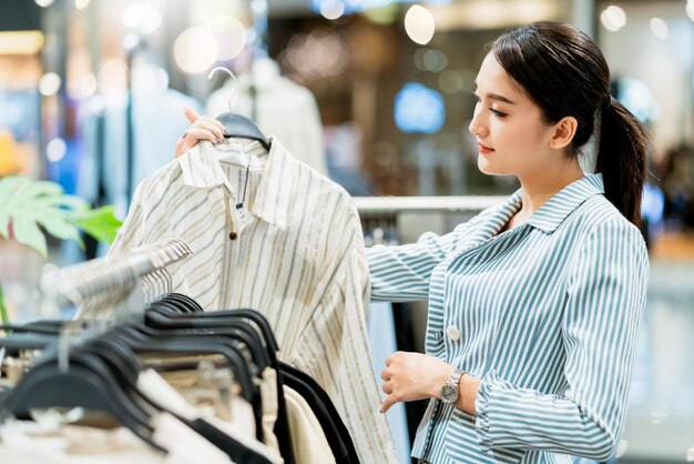 Mulher asiática inteligente e atraente gosta de escolher um pano de vestido adequado com rack de pano no fundo do shopping boutique