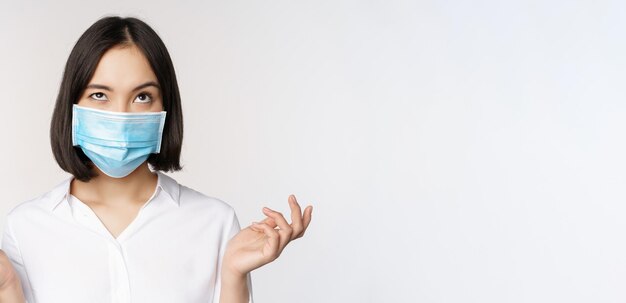 Mulher asiática irritada com máscara facial médica encolhendo os ombros e olhando para cima com expressão de rosto incomodado em pé sobre fundo branco