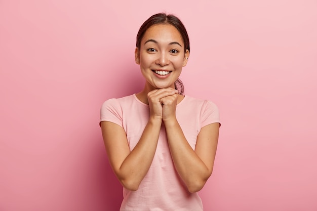 Mulher asiática jovem feliz com cabelo escuro penteado, mantém as mãos juntas sob o queixo, feliz em ouvir boas notícias, tem uma aparência charmosa, usa uma camiseta rosa casual, expressa boas emoções, tem piercing na orelha