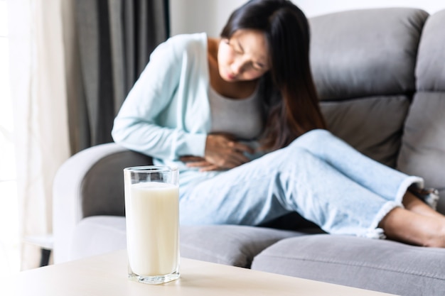 Mulher asiática jovem infeliz tendo uma forte dor de estômago com um copo de leite na mesa. intolerância à lactose, conceito de alergia alimentar.