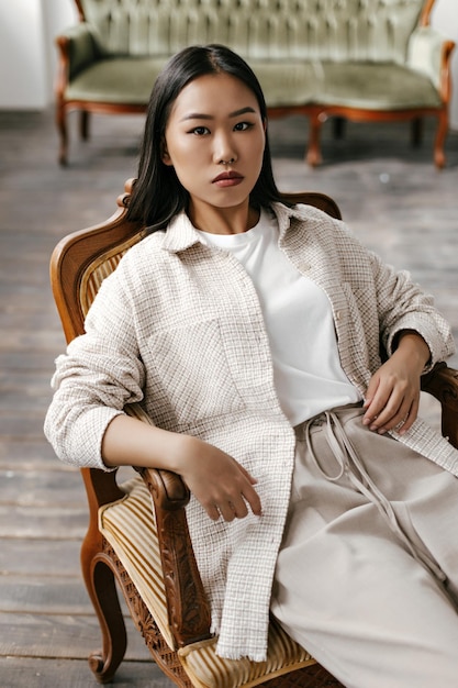 Mulher asiática morena de olhos castanhos em calças bege, camiseta branca e cardigã estiloso, sentado em uma poltrona clássica listrada macia