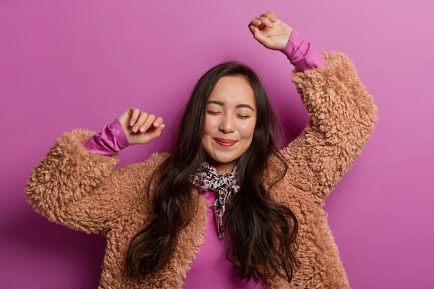 Mulher asiática morena feliz dança, levanta as mãos no ar, gosta de música, faz movimentos legais, convida para dançar com ela, tem clima romântico, olhar terno