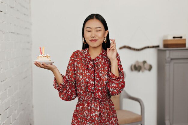 Mulher asiática morena feliz em um vestido floral fazendo um pedido e segurando um pedaço saboroso de bolo de aniversário