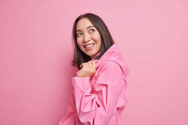 Mulher asiática morena muito feliz fica meio virada contra a parede rosa tem bom humor usa jaqueta elegante com capuz pensa em algo agradável poses feliz interior. Conceito de emoções