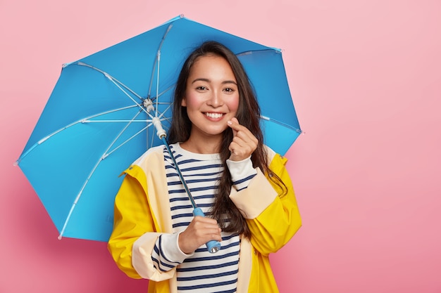 Mulher asiática muito carinhosa faz um signo coreano, tem uma expressão feliz, um sorriso gentil, fica sob o guarda-chuva, usa uma capa de chuva amarela