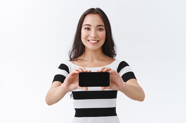 Mulher asiática muito charmosa em uma camiseta listrada, segurando o smartphone horizontalmente e sorrindo ao apresentar o jogo ou aplicativo, recomendar um bom aplicativo para baixar e usar, ficar com o fundo branco