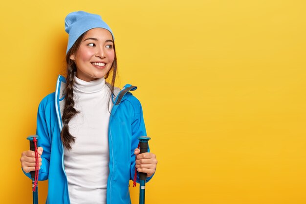 Mulher asiática muito sorridente gosta de caminhadas escandinavas, faz caminhada, olha de lado, penteava trança, usa chapéu e jaqueta azuis, segura bastões de trekking, isolada na parede amarela