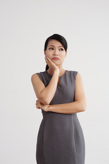 Mulher asiática no vestido posando no estúdio, segurando a mão na bochecha com expressão de rosto pensativo