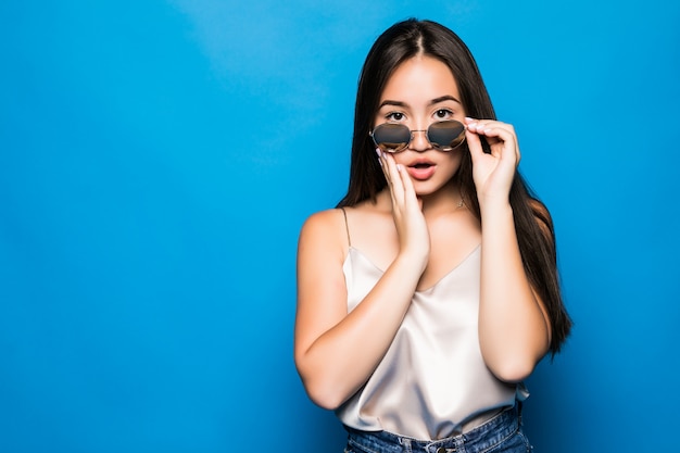 Mulher asiática nova com pose de surpresa isolada sobre fundo azul. retrato de uma linda mulher asiática no chapéu de palha e óculos escuros sobre fundo azul